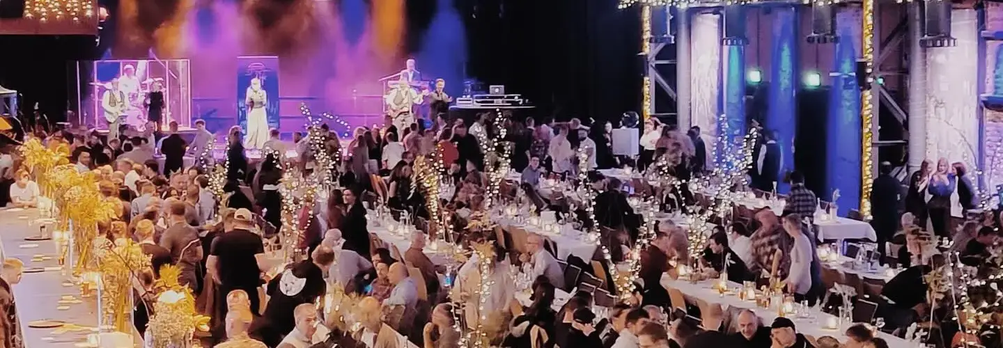 Eine große Indoor-Veranstaltung mit vielen Menschen, die an langen, dekorierten Tischen unter Lichterketten sitzen; eine Bühne mit Darstellern und violetter Beleuchtung ist am anderen Ende des Veranstaltungsortes zu sehen.