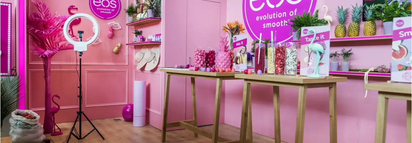 eventberatung-duisburg Ein pinkfarbener Messestand mit eos-Branding, Holztischen mit Produktboxen und Dekorationen, einer Ringleuchte auf einem Ständer, Pflanzen und Regalen mit farbenfrohen Artikeln vor einer pinkfarbenen Wand.