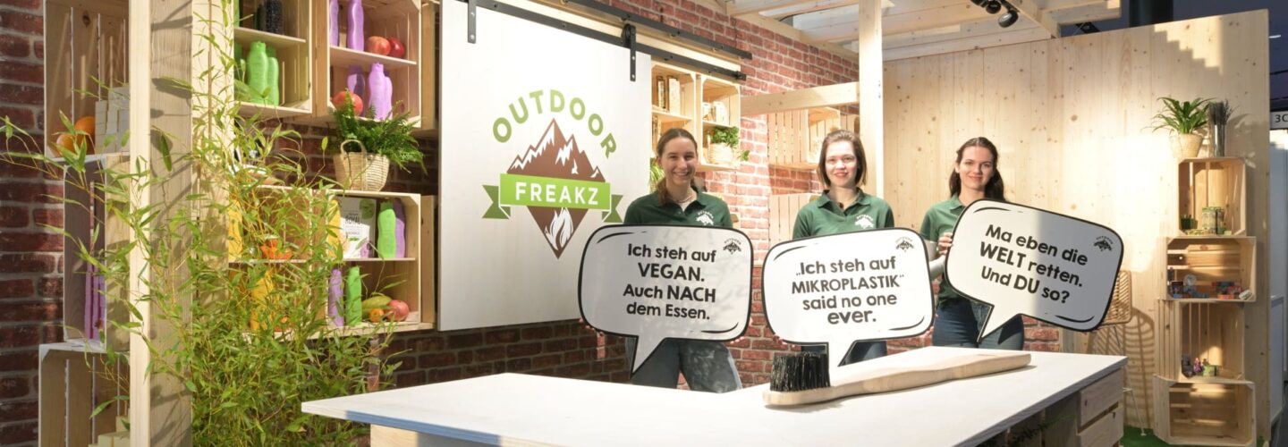 Drei Personen stehen hinter einem Holztresen in einem Stand mit der Aufschrift Outdoor Freakz und halten deutsche Sprechblasen. Der Stand ist mit Holzregalen, Grünpflanzen und verschiedenen Flaschen ausgestattet. Die Atmosphäre ist hell und aufgeräumt.