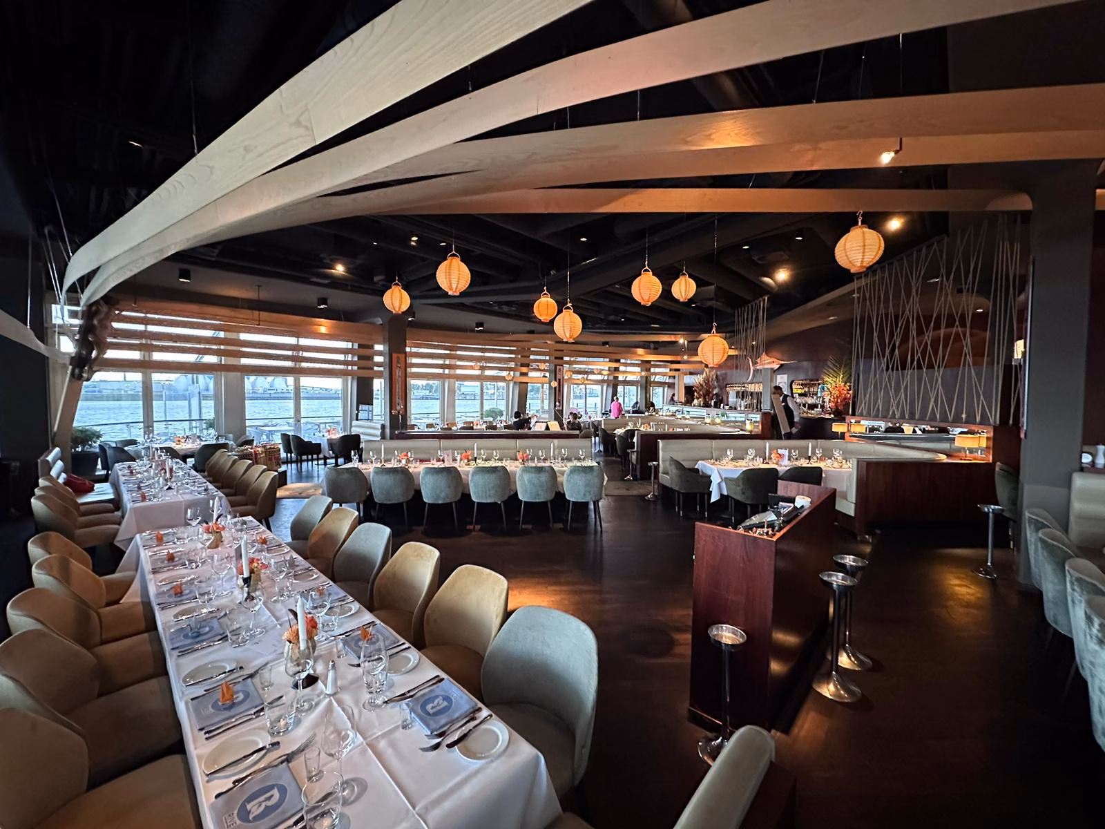 Ein modernes Restaurant mit langen Tischen zum Essen, gepolsterten Stühlen, Hängelampen und einem Barbereich im Hintergrund. Große Fenster säumen eine Wand und lassen natürliches Licht herein.