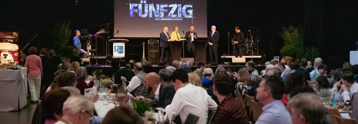 eventberatung-greifswald Eine große Gruppe von Menschen sitzt an geschmückten Tischen in einer Halle und blickt auf eine gut beleuchtete Bühne, auf der mehrere Personen neben einem Podium und einem Bildschirm stehen, auf dem das Wort FÜNFZIG erscheint.