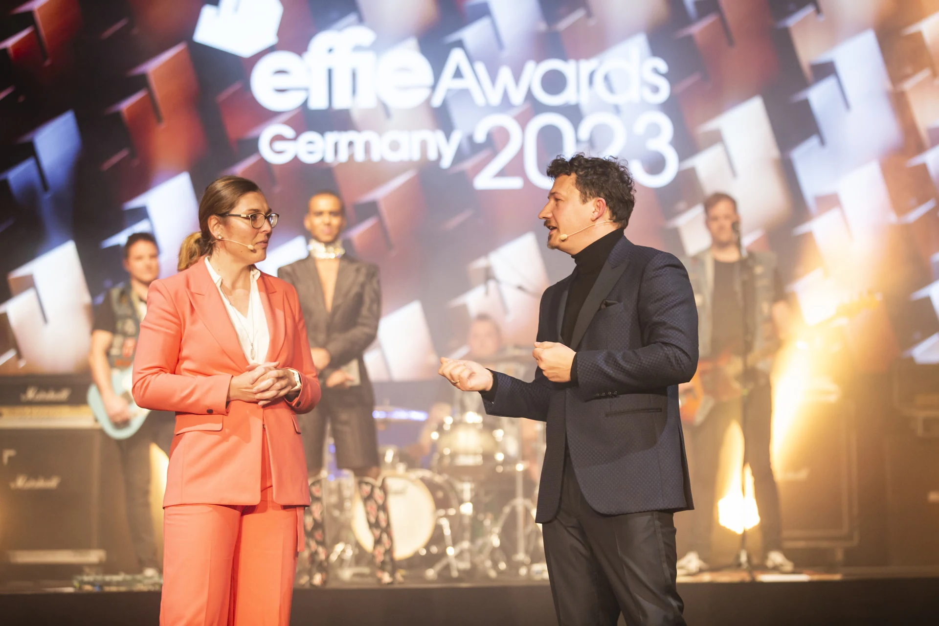 Zwei Personen sprechen auf der Bühne der Veranstaltung Effie Awards Germany 2023. Im Hintergrund ist eine Band mit Schlagzeug und Gitarren zu sehen, hinter ihnen wird das Veranstaltungslogo projiziert.