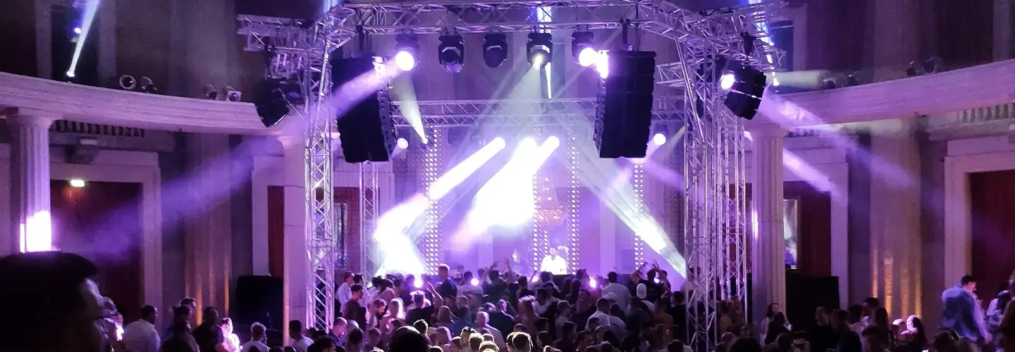 Ein überfülltes Konzert oder eine Party in einer Halle mit Menschen, die auf eine Bühne blicken, die von lila und weißen Scheinwerfern beleuchtet wird. Die Bühne hat einen Metallrahmen, große Lautsprecher und Lichtstrahlen, die auf das Publikum scheinen.