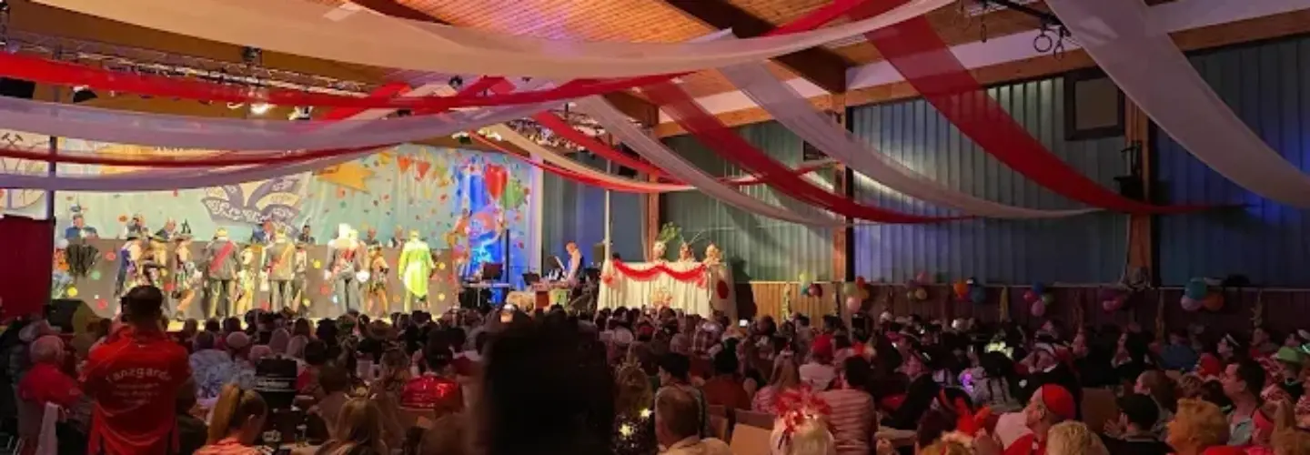 eventberatung-schwerin Eine große Menschenmenge sitzt an Tischen in einem geschmückten Saal und beobachtet eine Bühnenaufführung. Rote und weiße Luftschlangen hängen von der Decke. Viele Menschen sind rot gekleidet, und die Atmosphäre wirkt festlich und lebendig.