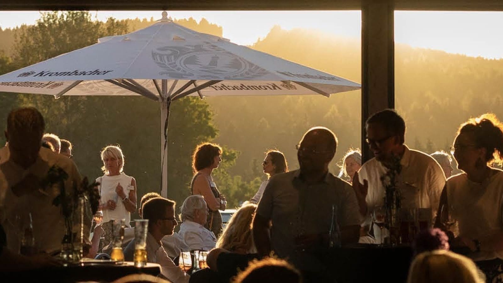 Menschen versammeln sich bei Sonnenuntergang im Freien unter großen Sonnenschirmen, mit Hügeln und Bäumen im Hintergrund. Es scheint sich um ein gesellschaftliches Ereignis oder eine Party zu handeln, bei der die Gäste zusammen stehen und sitzen und sich unterhalten.
