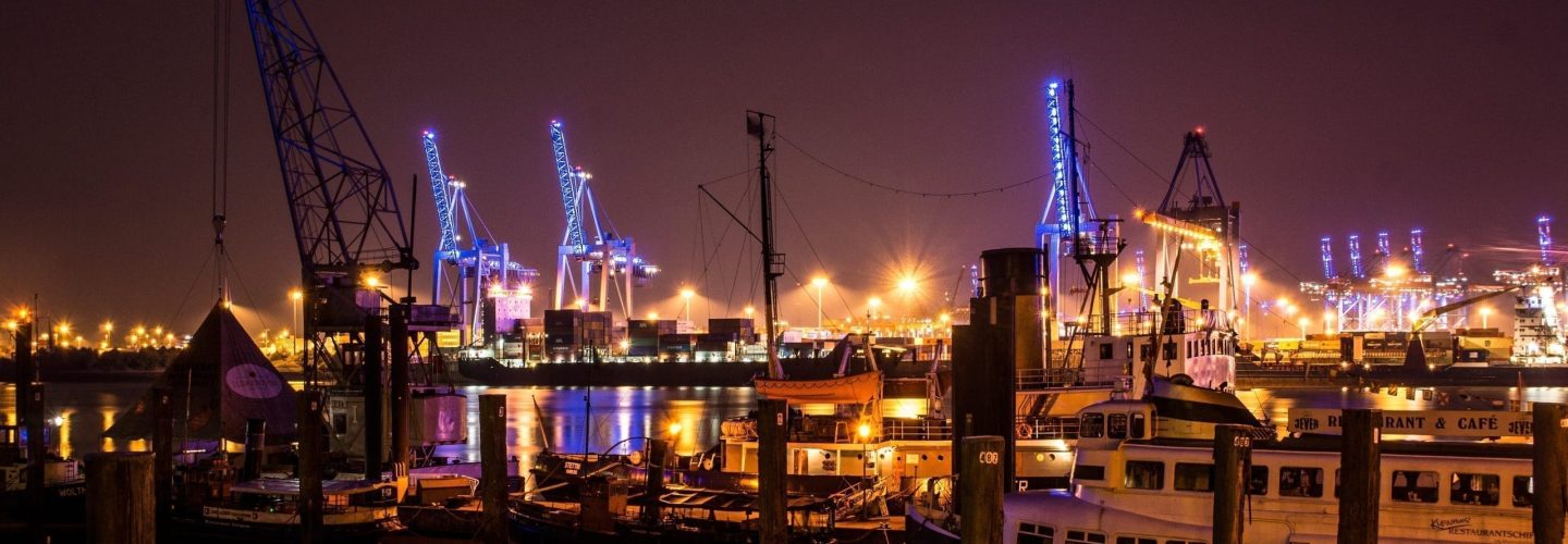 hamburg-1118753 Ein Hafen bei Nacht mit beleuchteten Kränen, angedockten Booten und Schiffen, die sich im Wasser spiegeln. Helle Lichter heben die Industriestrukturen hervor und verleihen der Szene ein buntes Leuchten vor dem dunklen Himmel.