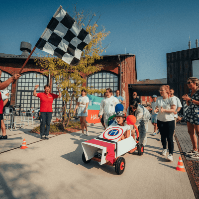 Eine Gruppe von Menschen sieht zu, wie eine Person in einem selbst gebauten Rennwagen, der mit einem roten und blauen Muster verziert ist, die Ziellinie überquert. Ein Beamter schwenkt die Zielflagge. Die Veranstaltung findet an einem sonnigen Tag im Freien statt.