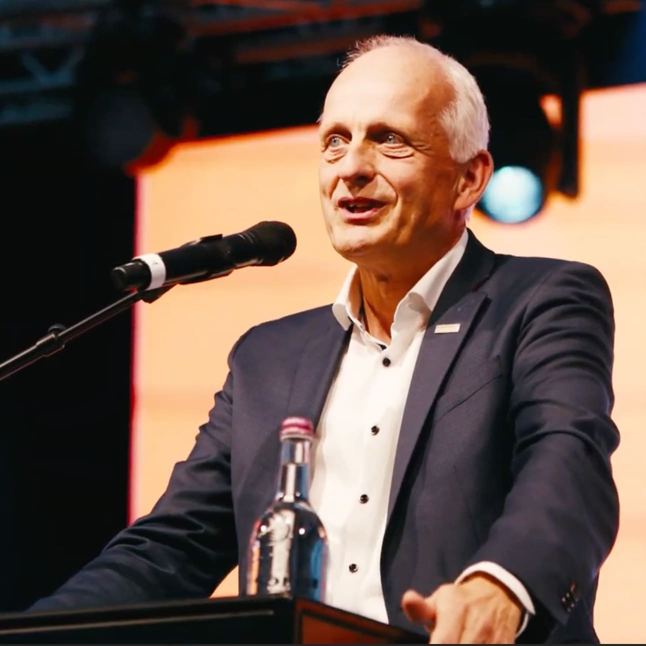Ein Mann im Anzug steht an einem Podium und spricht in ein Mikrofon, vor dem er eine Flasche Wasser hält. Im Hintergrund sind helle, warme Beleuchtung und unscharfe Bühnenelemente zu sehen.