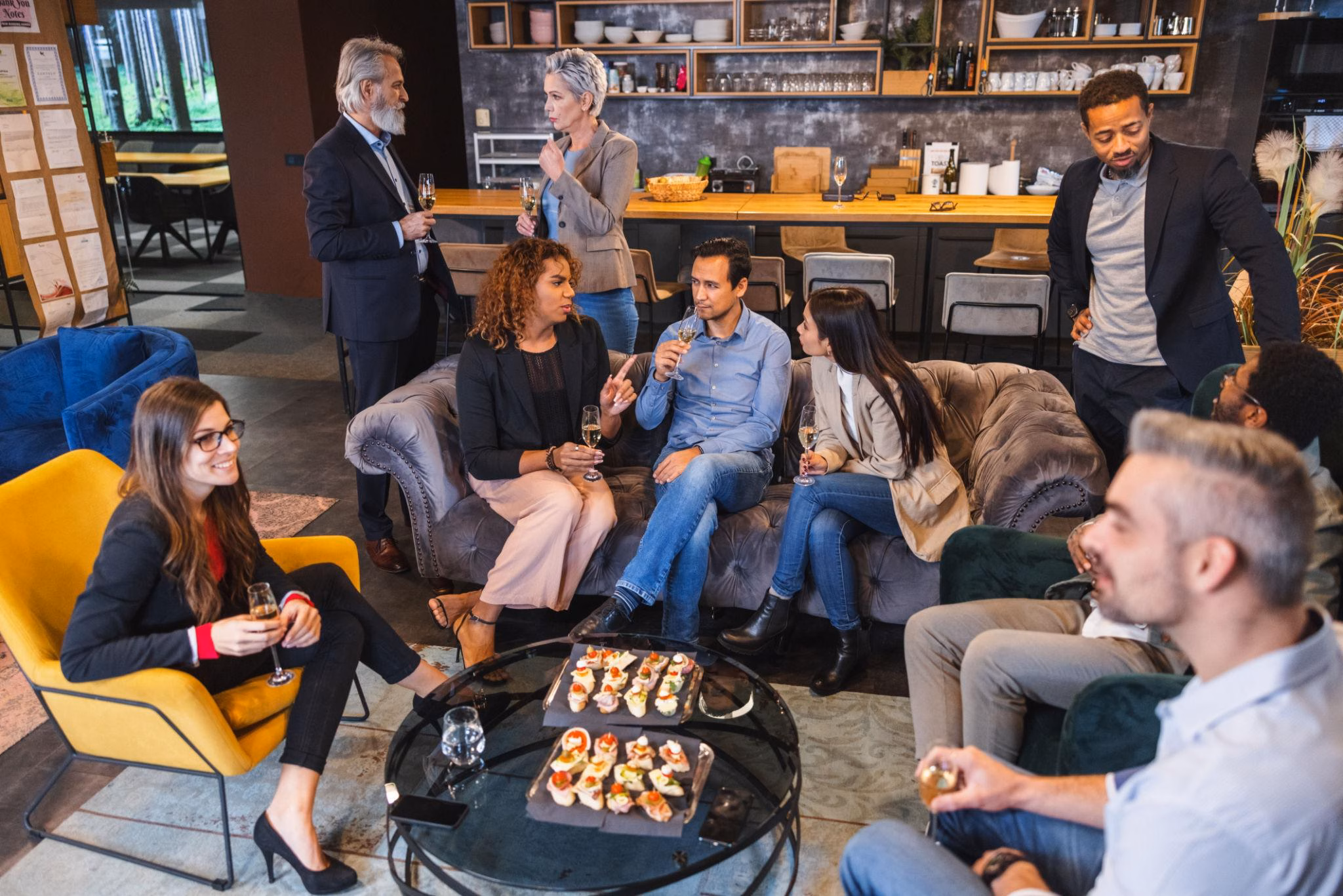 Eine Gruppe von Personen in Geschäftskleidung sitzt und steht in einem modernen Lounge-Bereich, hält Getränke in der Hand und unterhält sich. Auf einem gläsernen Couchtisch in der Mitte der Gruppe sind kleine Häppchen angerichtet.
