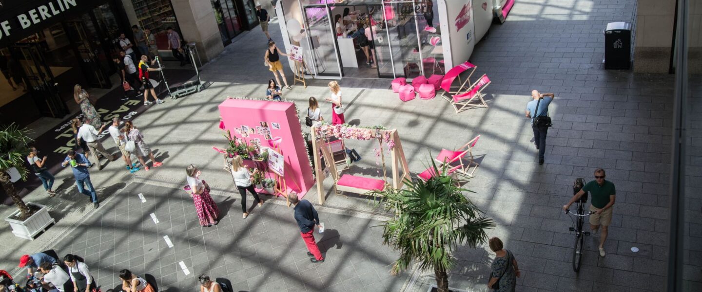 Ein überdachtes Einkaufszentrum mit Menschen, die gehen und sitzen. In der Mitte ist ein rosa-weißer Werbestand mit Displays und Sitzgelegenheiten aufgebaut, umgeben von Palmen und unter einer Glasdecke, die Sonnenlicht hereinlässt.