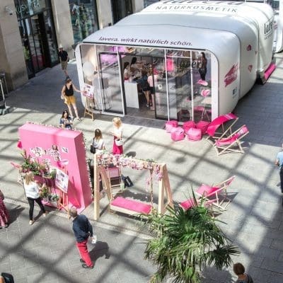 Eine Szene in einem Einkaufszentrum mit einem weißen aufblasbaren Stand und mehreren Ausstellungsbereichen mit rosafarbenen Themen. Die Menschen gehen, sitzen auf Liegestühlen und interagieren mit den Ausstellungsstücken. Natürliches Licht strömt durch das Dach.