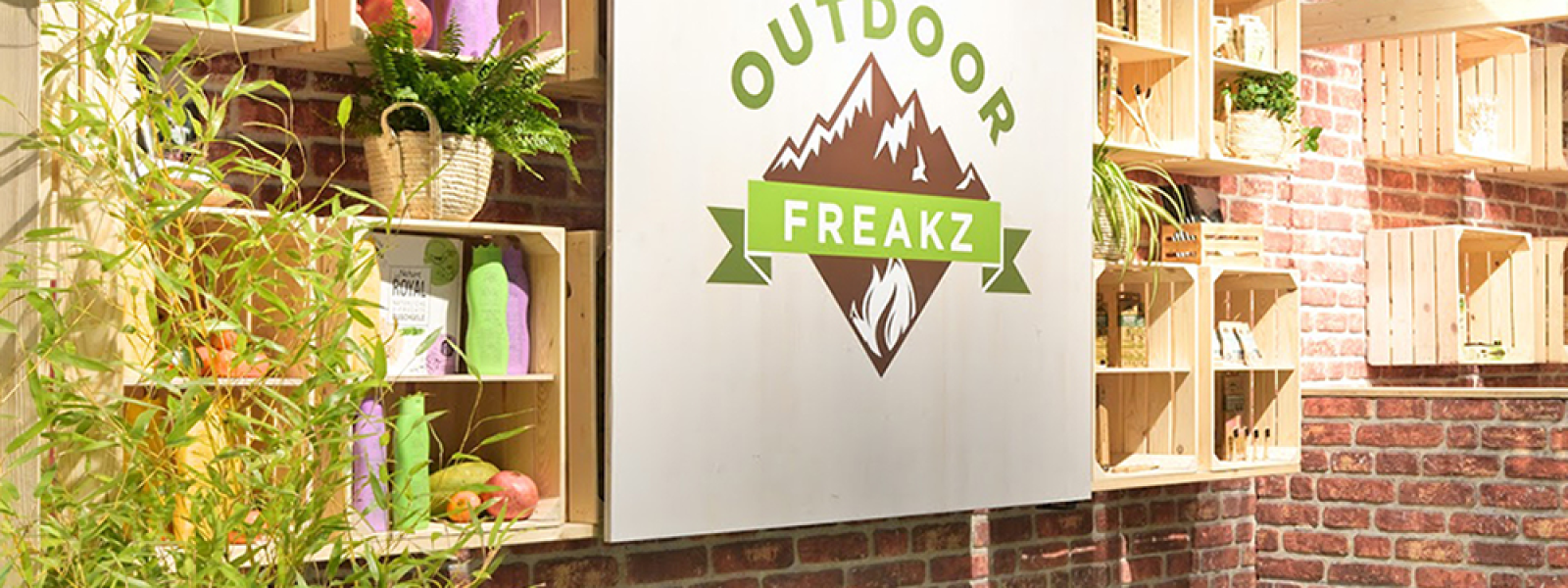 Ein Ladeninneres mit einem Schild "Outdoor Freakz" auf einer weißen Tafel. In den Holzregalen stehen bunte Flaschen und Pflanzen, und die Wand besteht aus Sichtziegeln. Im Vordergrund stehen eine weiße Theke und etwas Bambus.