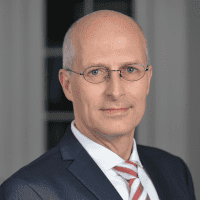 Ein Mann mittleren Alters mit Brille, kurzen grauen Haaren und glatt rasiertem Gesicht trägt einen dunklen Anzug, ein weißes Hemd und eine gestreifte Krawatte und posiert vor einem unscharfen Innenhintergrund.