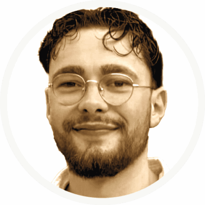 unnamed-1 Ein Mann mit kurzen dunklen Haaren, einer Brille und einem Bart lächelt leicht. Er ist von den Schultern aufwärts vor einem schlichten weißen Hintergrund abgebildet.