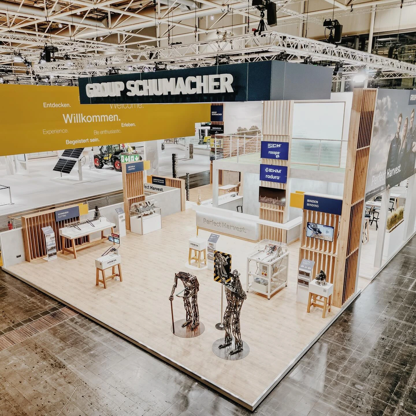 Ein geräumiger Messestand der Schumacher-Gruppe mit Holzakzenten, Informationsdisplays, Sitzgelegenheiten, Produktvitrinen und Landmaschinenmodellen aus Metall auf einem polierten Boden in einer großen Messehalle.