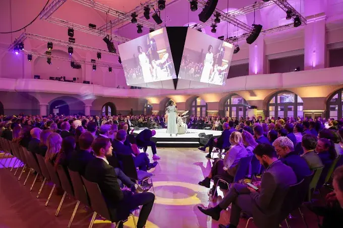 Ein großes Publikum sitzt in Reihen um eine kreisförmige Bühne herum und beobachtet einen Redner bei hellem Licht. Die Veranstaltung findet in einer geräumigen, modernen Halle mit großen Bildschirmen statt, auf denen die Bühne zu sehen ist.