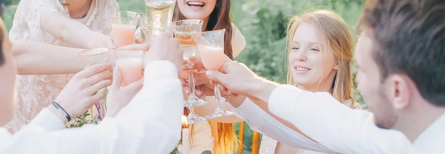 veranstaltungsagentur-lueneburg Sechs Personen sitzen um einen Tisch im Freien und stoßen mit ihren Gläsern an. Der Tisch ist mit Blumen, Kerzen und Tellern mit Speisen gedeckt. Die Umgebung wirkt hell und natürlich, mit viel Grün und Bäumen im Hintergrund.