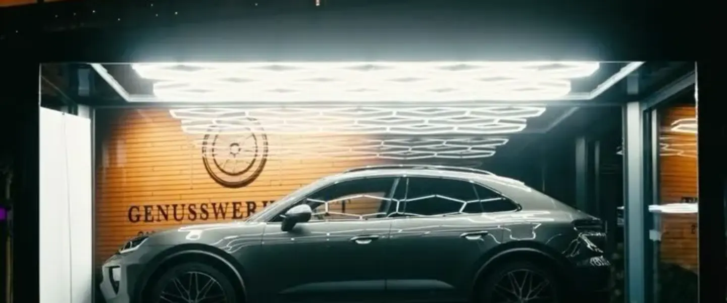 Ein dunkelgraues Auto ist in einem hell erleuchteten Glasgehäuse mit Holzwänden und dem Wort GENUSSWERK im Hintergrund ausgestellt. Geometrische Deckenleuchten erhellen die Szene.
