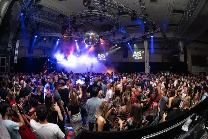 veranstaltungsagentur-schwerin Eine große Menschenmenge versammelt sich bei einem Hallenkonzert oder einer Party mit bunten Bühnenlichtern, einer Discokugel über dem Kopf und einem DJ-Pult im Hintergrund, das "ASS Radio" auf Bildschirmen zeigt.