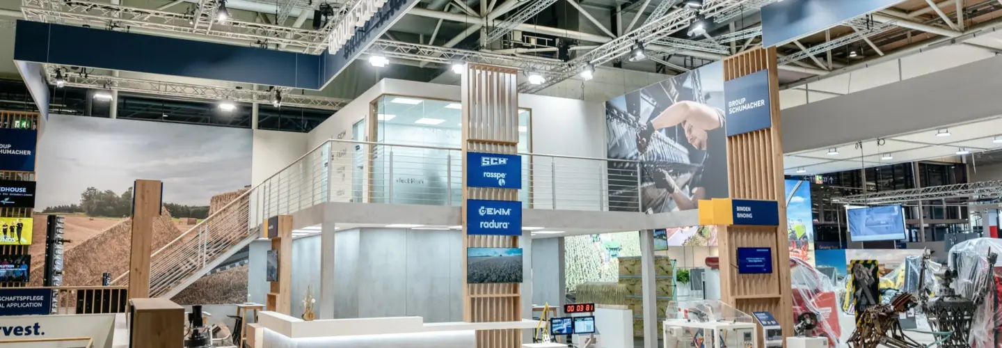 veranstaltungsmanagement-bochum Ein moderner Messestand für die Schumacher-Gruppe ist mit einem Empfangstresen, Hockern, Produktdisplays und Werbebannern unter heller Beleuchtung ausgestattet. Eine Treppe führt zu einer oberen Ebene mit verschiedenen landwirtschaftlichen Themen.