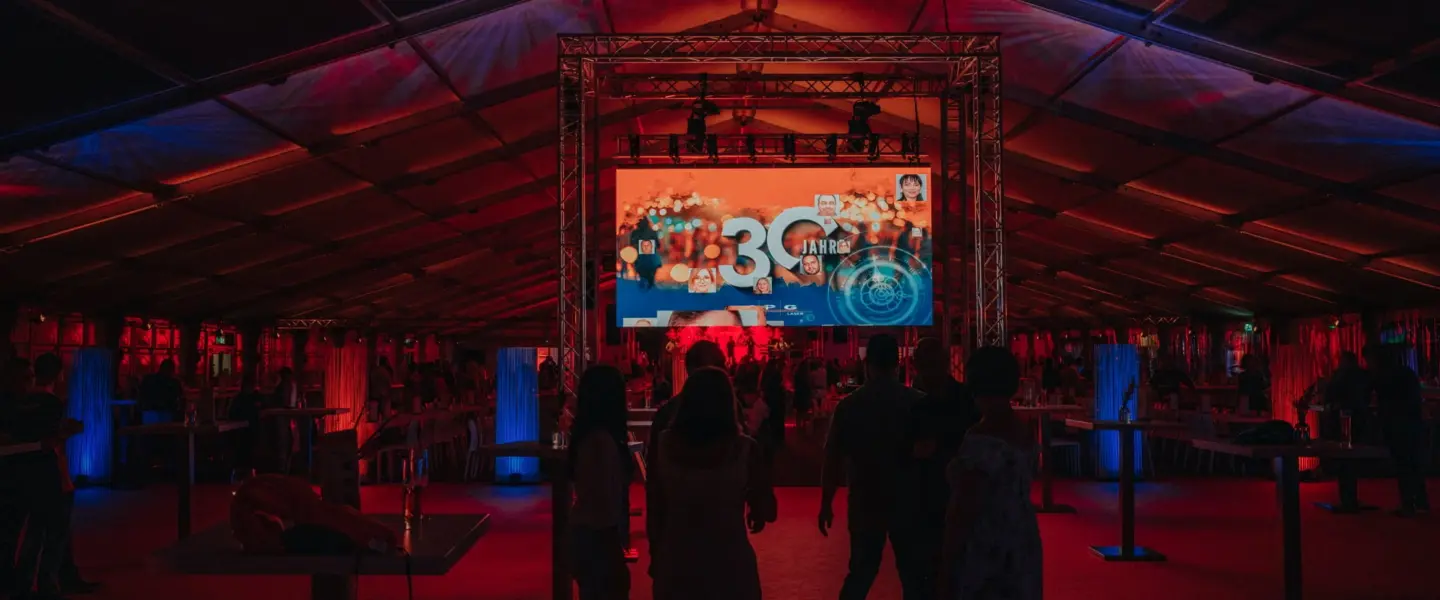 Die Menschen stehen in einem großen Zelt mit roter und blauer Beleuchtung und blicken auf eine große Leinwand, auf der bunte Grafiken und die Zahl 30 zu sehen sind. An den Seiten des Zeltes sind Tische und Stühle aufgestellt.