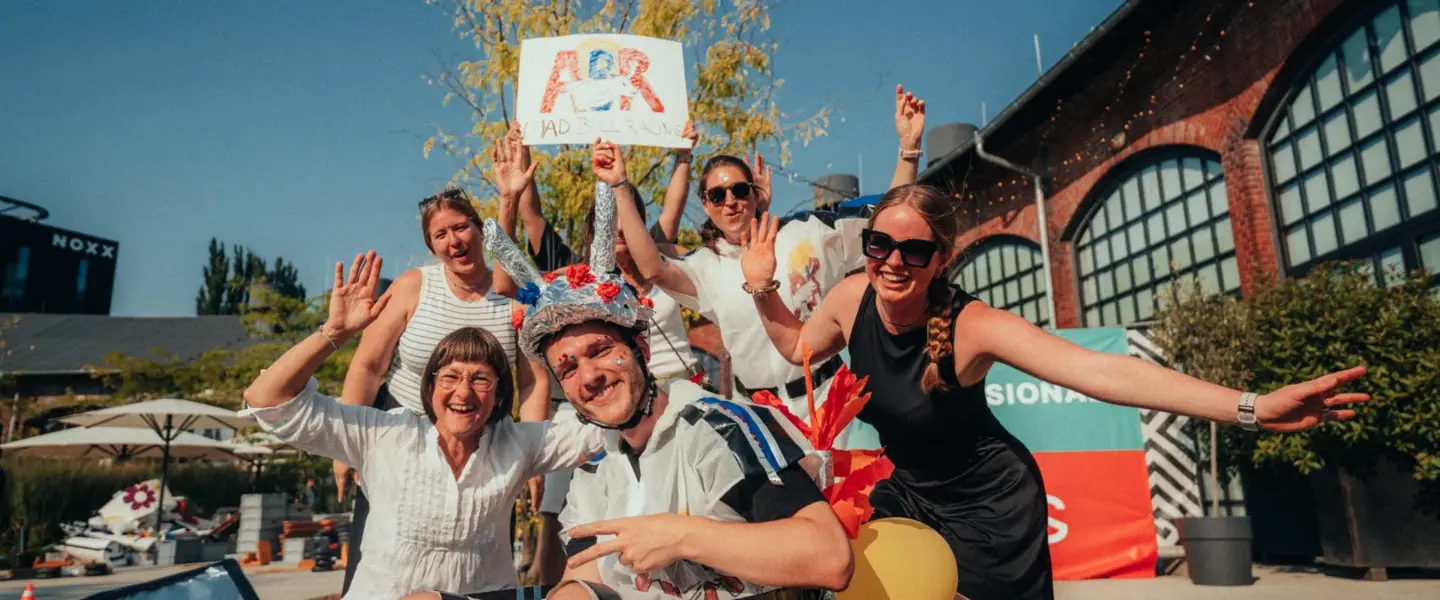 veranstaltungsmanagement-greifswald Eine Gruppe von sechs Erwachsenen posiert im Freien mit einem selbstgebauten Gokart. Eine Person sitzt in dem Kart und trägt einen Helm, während die anderen lächelnd und winkend herumstehen. Ein Schild mit der Aufschrift "ADR" wird hochgehalten. Im Hintergrund ist ein Industriegebäude zu sehen.