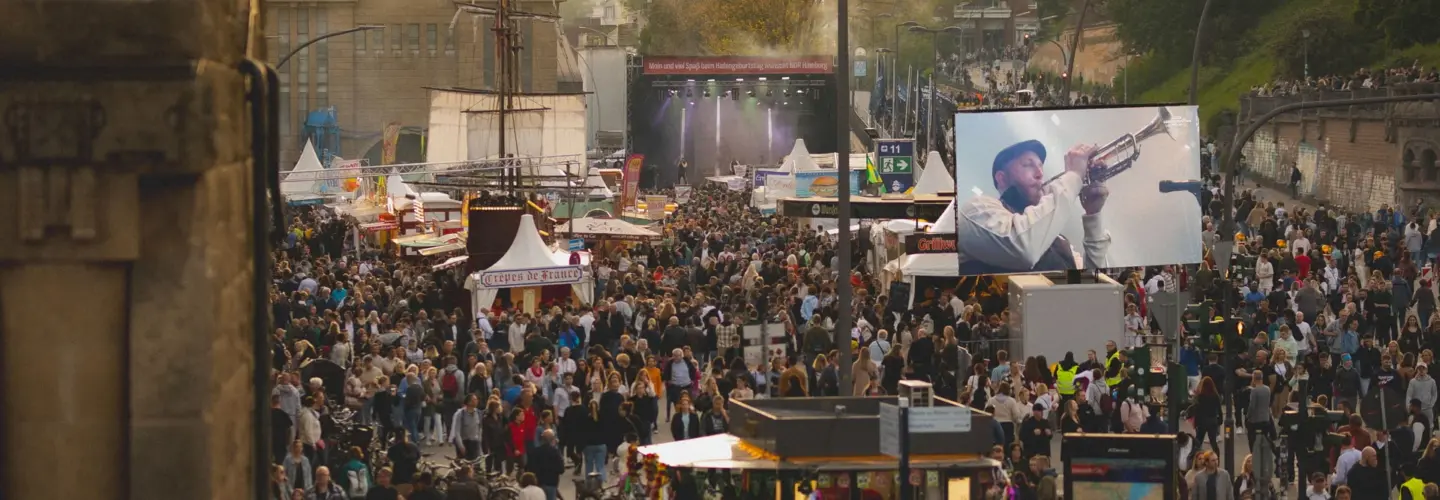 Eine große Menschenmenge versammelt sich auf einem Fest im Freien mit Essensständen, Bühnenauftritten und einem großen Bildschirm, auf dem ein Musiker Trompete spielt; im Hintergrund sind städtische Gebäude und Bäume zu sehen.