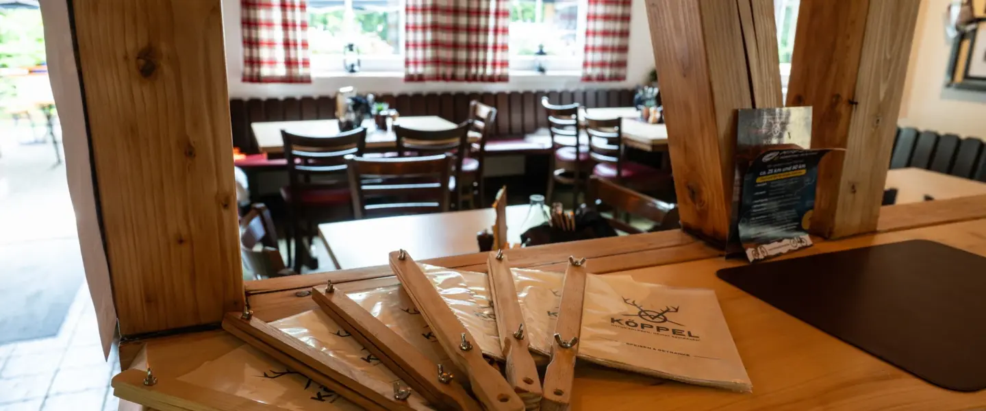 veranstaltungsmanagement-lueneburg Ein hölzerner Tresen mit mehreren hölzernen Menütafeln und Papiermenüs mit der Aufschrift Köppel. Im Hintergrund sind Tische und Stühle in einem Restaurant mit rot-weiß-karierten Vorhängen aufgestellt.