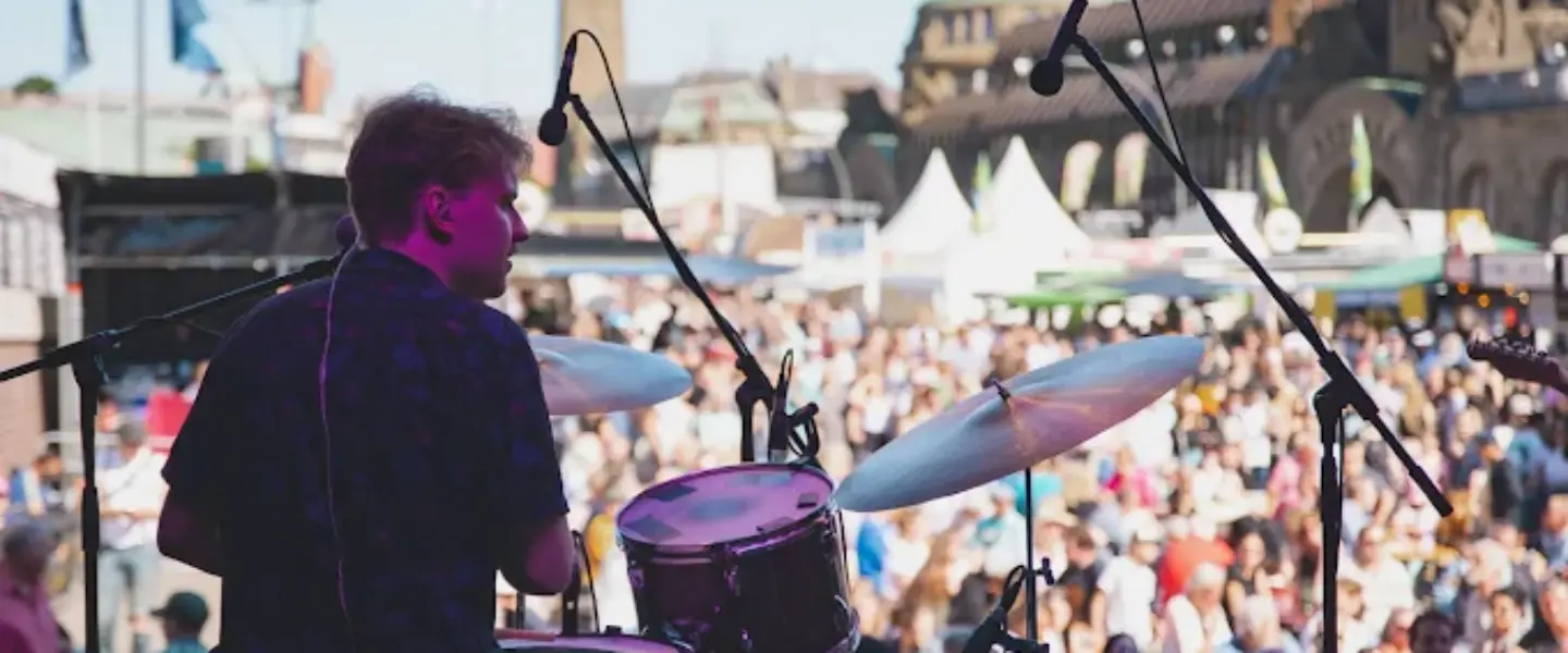 veranstaltungsmanagement-schwerin Ein Schlagzeuger spielt auf der Bühne eines Konzerts im Freien vor einer großen Menschenmenge. Mikrofone, Schlagzeug und Festivalzelte sind zu sehen, mit historischen Gebäuden und einem klaren blauen Himmel im Hintergrund.